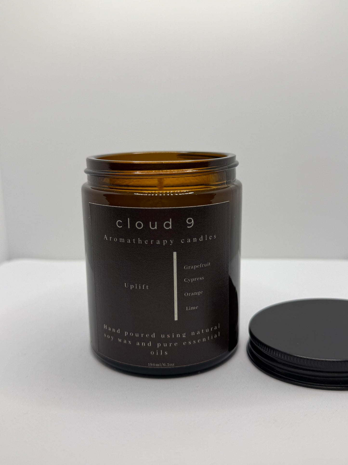 Amber glass jar candle labeled ‘Uplift’, featuring grapefruit, cypress, orange, and lime essential oils—set on a neutral-toned surface beside a potted plant, with soft overhead lighting.