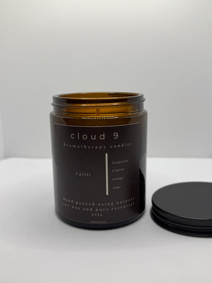 Amber glass jar candle labeled ‘Uplift’, featuring grapefruit, cypress, orange, and lime essential oils—set on a neutral-toned surface beside a potted plant, with soft overhead lighting.