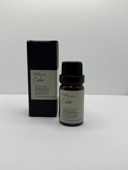 Close-up of a hand-poured ‘Calm’ essential oil blend bottle beside fresh ylang ylang and marjoram blooms, suggesting a soothing aromatherapy ritual.