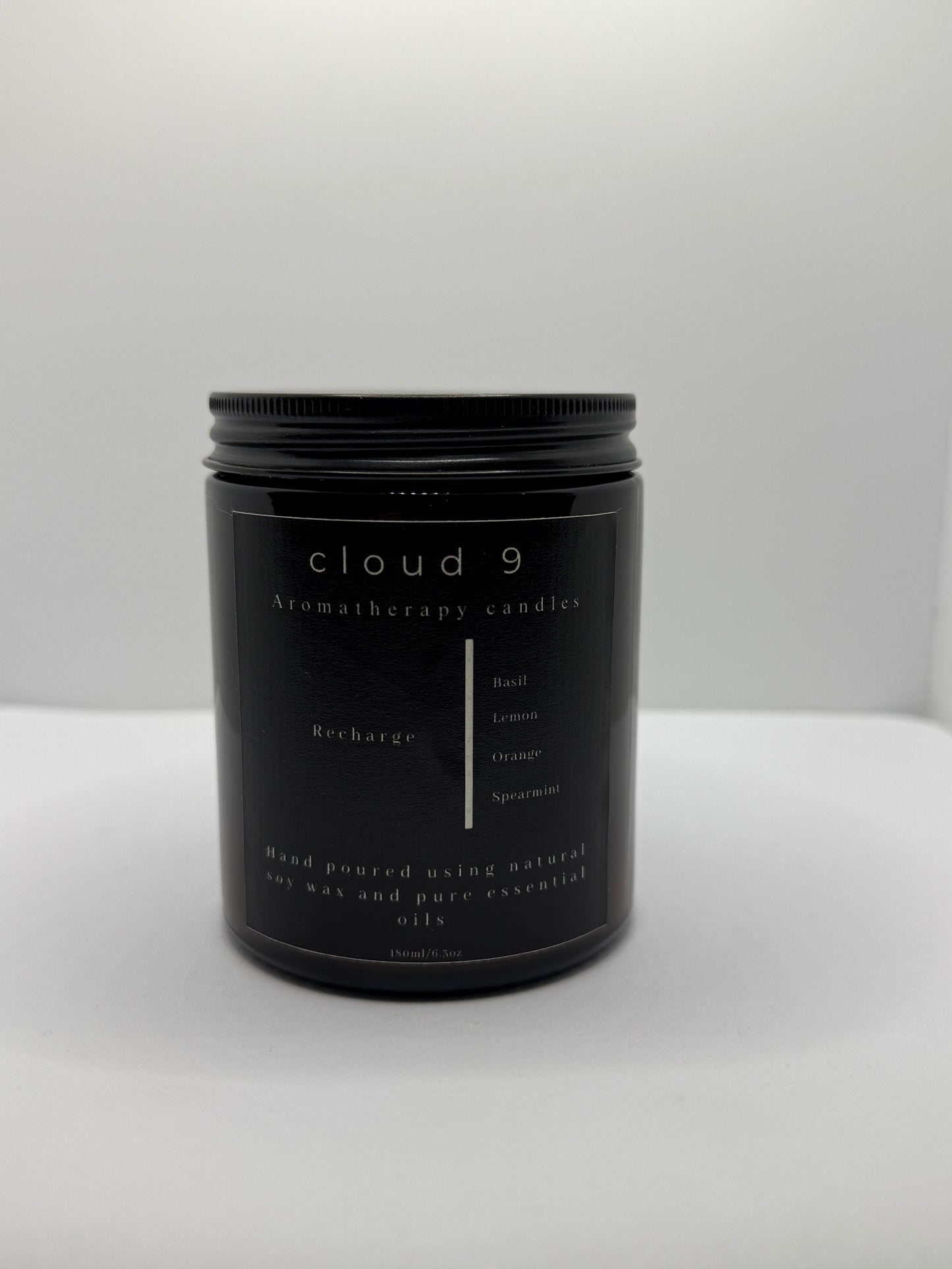 A sleek, dark amber glass jar candle labeled ‘Recharge’ with a beige label, showcasing the citrus and herbal scent blend—lemon, basil, orange, and spearmint—in a clean, minimalist setting.