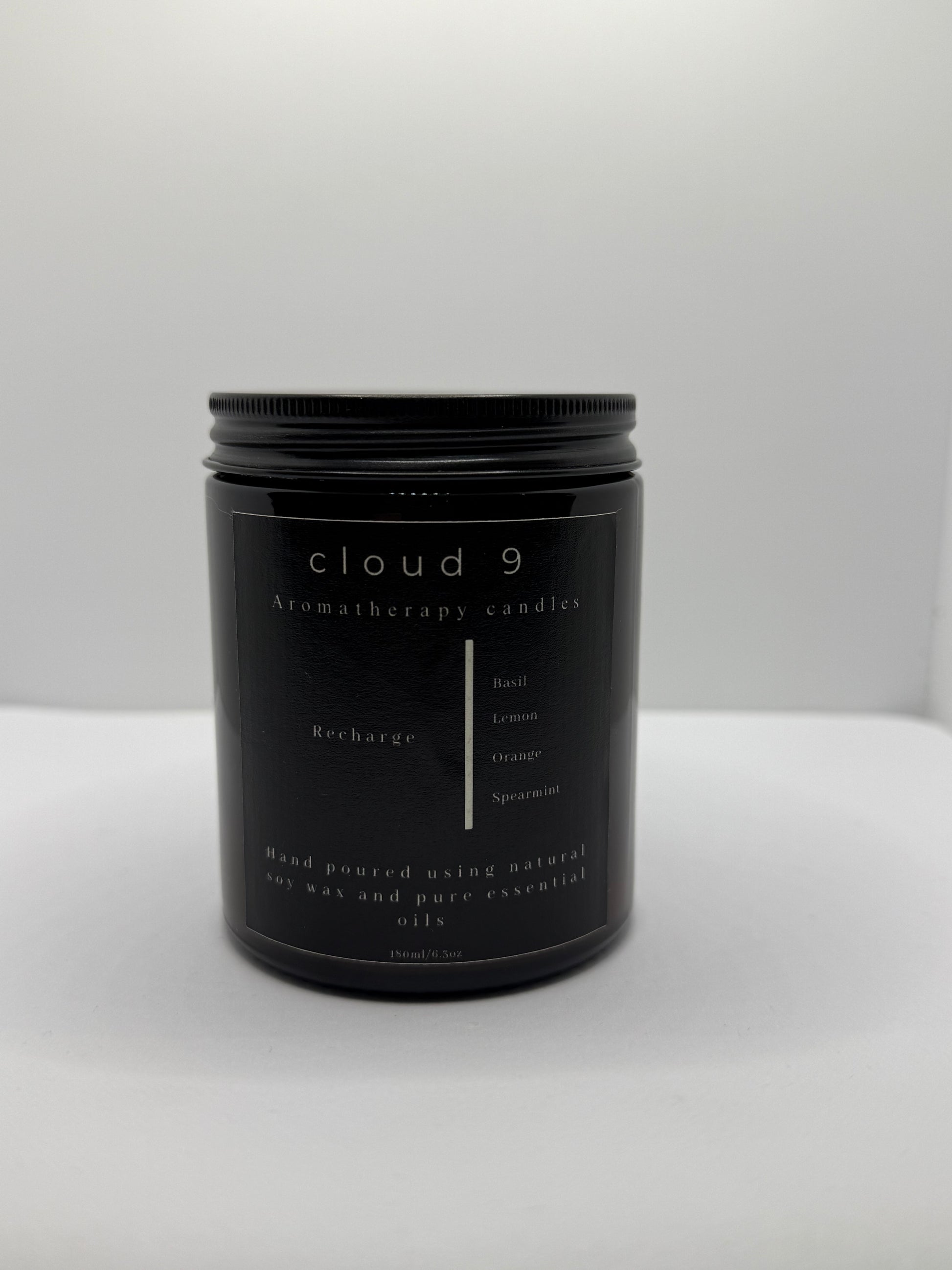 A sleek, dark amber glass jar candle labeled ‘Recharge’ with a beige label, showcasing the citrus and herbal scent blend—lemon, basil, orange, and spearmint—in a clean, minimalist setting.