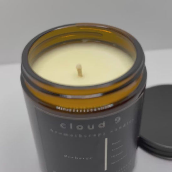 The ‘Recharge’ candle burning softly beside green foliage, evoking a serene and revitalizing atmosphere with its natural, mood-boosting essential oil blend.