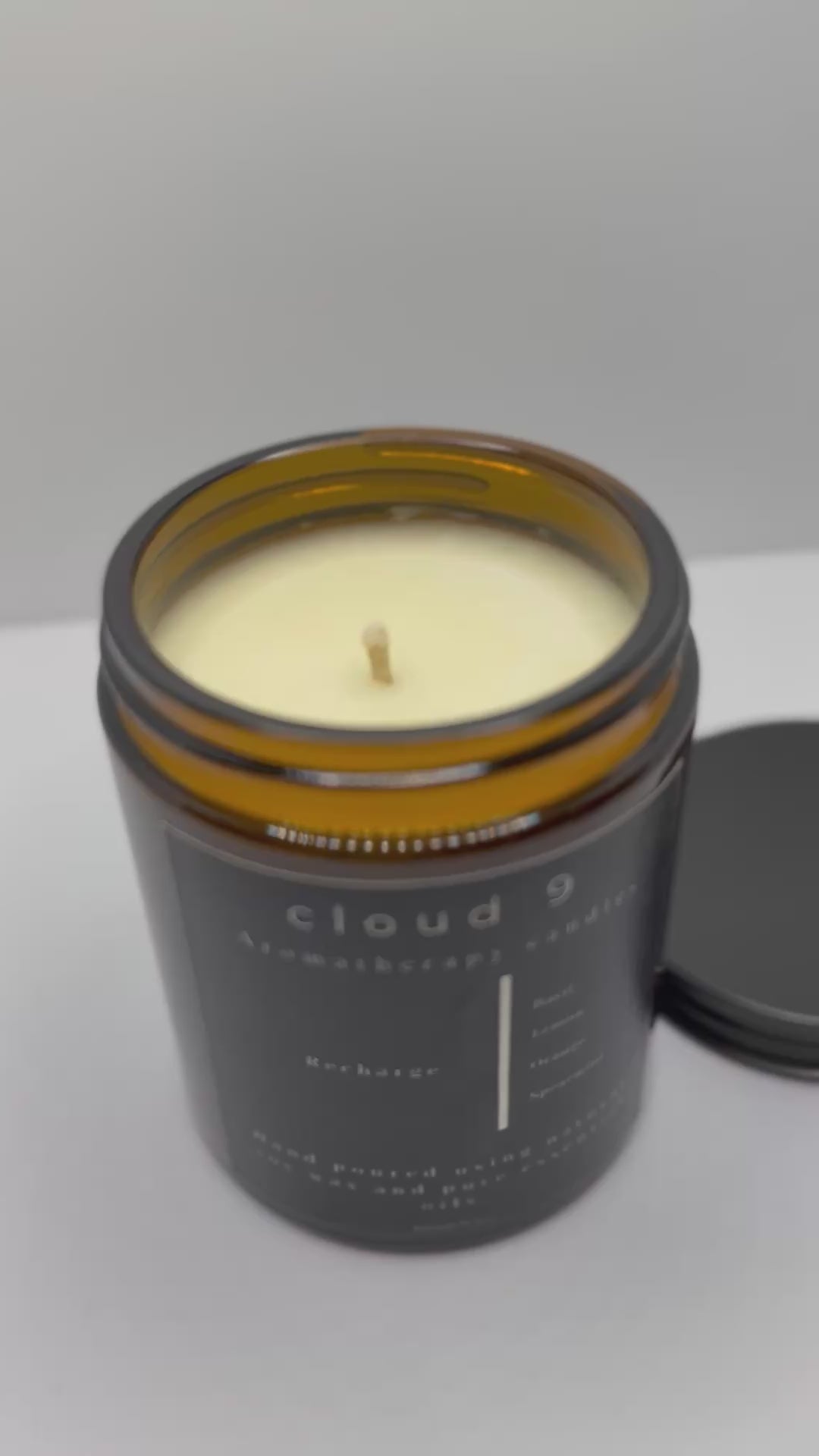 The ‘Recharge’ candle burning softly beside green foliage, evoking a serene and revitalizing atmosphere with its natural, mood-boosting essential oil blend.